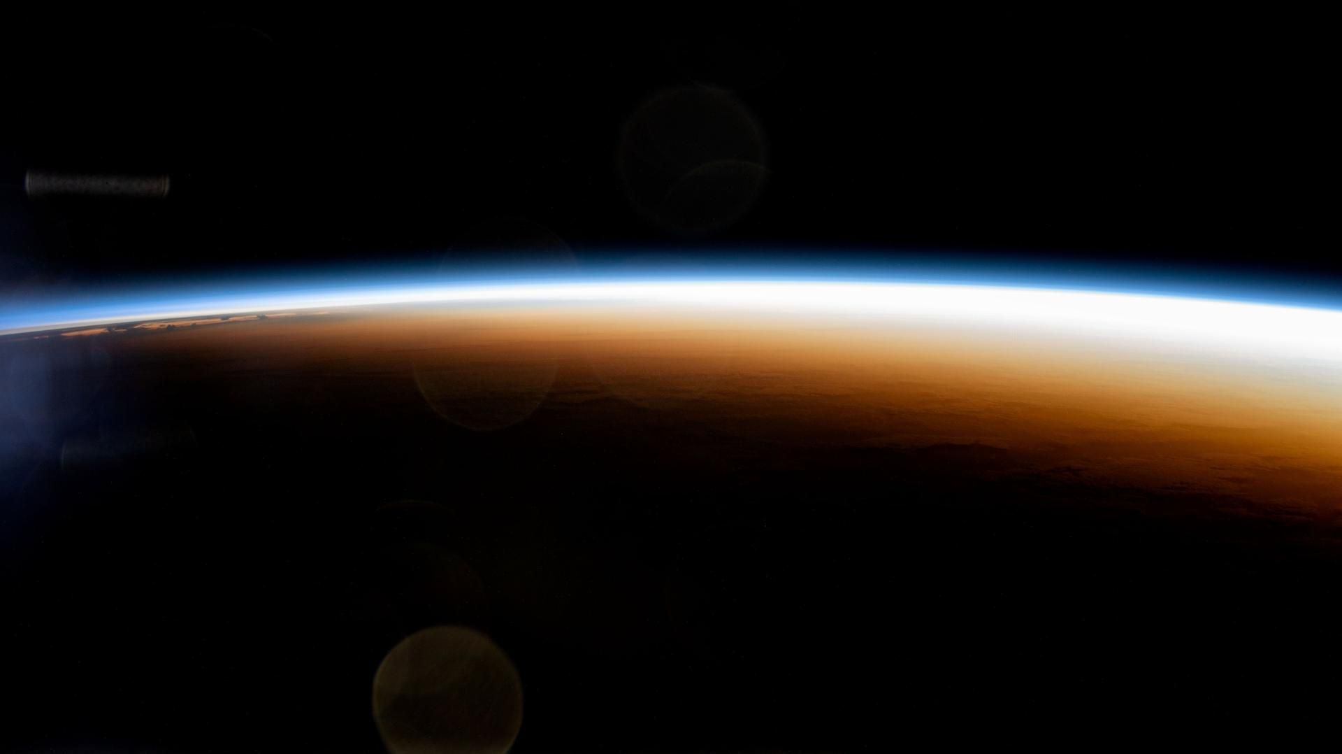 An orbital sunrise seen from the International Space Station: a thin blue line of atmosphere above an orange horizon.